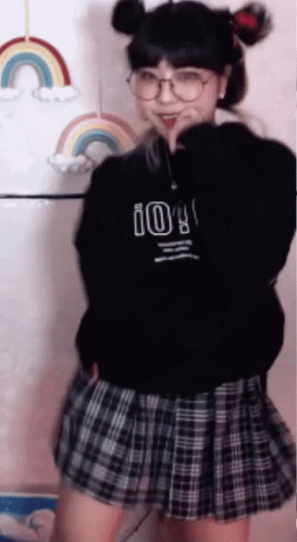 a girl wearing a black sweater and a plaid skirt is dancing in front of a rainbow .