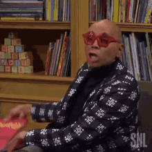 a bald man wearing red sunglasses and a black jacket is sitting in front of a bookshelf .
