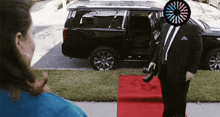 a man in a suit stands on a red carpet in front of a suv
