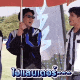 a man wearing sunglasses is standing under an umbrella while another man stands behind him .