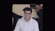 a man wearing glasses and a white hoodie is smiling while sitting in a chair .