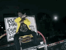 a man in a yellow jacket is sitting on a fence with a sign that says ku mal