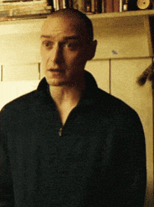 a man with a shaved head is standing in front of a bookshelf