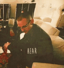 a man wearing glasses sits on a couch with the word rear written on his back