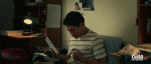 a man sits on a couch reading a piece of paper with kahar written on the bottom right