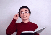 a woman wearing glasses and a red sweater is holding a book and smiling .