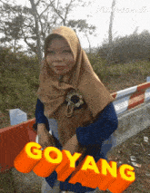 a woman in a hijab is standing in front of a red and white barrier with the word goyang written in yellow