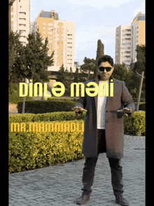 a man in a coat and sunglasses stands in front of a bush with the words dinle mani