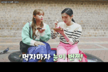 two women are sitting on a bench eating food in front of a brick wall with honeycam.org written on the bottom right