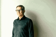 an older man wearing glasses and a black shirt stands in front of a white wall