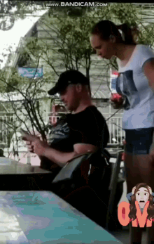 a man sits at a table looking at his phone and a woman stands behind him