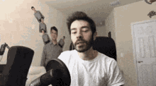 a man with a beard is sitting in front of a microphone in a room with other people standing around him .