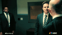 three men in suits and ties are standing in a dark room with #suitsla on the bottom right