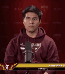 a man wearing a maroon hoodie that says sanguine on it stands in front of a microphone