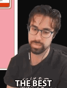 a man with a beard and glasses is sitting in front of a pink screen and says `` the best '' .