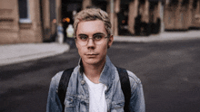 a young man wearing glasses and a denim jacket stands on a street