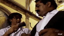 a man with a mustache is talking on a cell phone while a woman adjusts his tie .