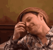 a man wearing a hat and a floral shirt is smiling while covering his face with his hand .