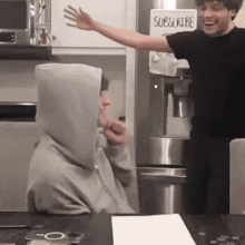 a man in a hoodie is standing next to another man in a black shirt in a kitchen .