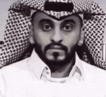 a man with a beard wearing a keffiyeh and a white shirt is looking at the camera .