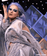 a drag queen with purple hair is wearing a gray and white dress