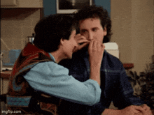 two men are sitting at a table and one of them is touching another man 's face
