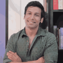 a man in a green shirt is smiling with his arms crossed in front of a bookshelf