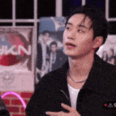 a young man wearing a black jacket and a necklace is talking to someone .