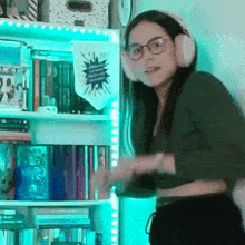 a woman wearing headphones and glasses is standing next to a bookshelf .