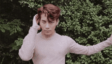 a man in a white sweater is standing in front of a bush and holding his hair .