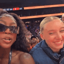 a woman wearing sunglasses stands next to another woman in front of a crowd at a stadium
