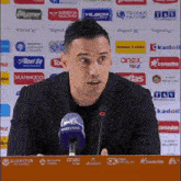 a man speaking into a microphone in front of a wall of advertisements including anex tour