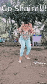 a woman in a floral shirt and blue pants is dancing in the dirt in front of a tree .