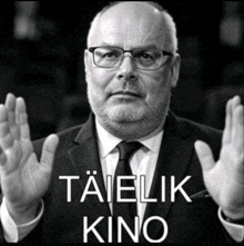 a man with glasses and a beard holds his hands up with the words taielik kino below him