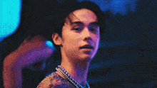 a man wearing a necklace of pearls looks at the camera with a woman in the background