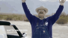 a man wearing overalls and a hat is standing in front of a car in the desert .