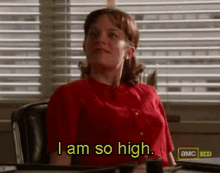 a woman in a red shirt is sitting at a desk saying i am so high
