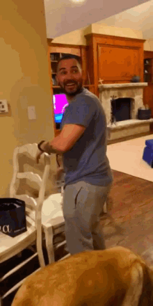 a man in a blue shirt stands in a living room next to a dog