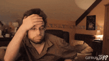 a man with glasses holds his hand to his forehead in a bedroom