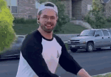 a man wearing a baseball cap and a white shirt is standing in front of a silver suv .