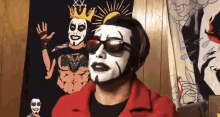 a man with white face paint and sunglasses is standing in front of a wall with a picture of a man in a crown .