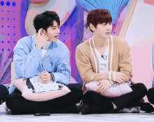 two young men are sitting next to each other on the floor and one of them is talking on the phone .
