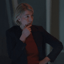 a woman wearing a black jacket and a red sweater has her hand on her chin