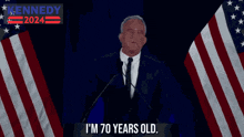 a man in a suit and tie is giving a speech in front of an american flag with the words kennedy 2024 behind him