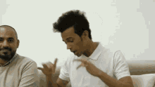 two men are sitting on a couch talking to each other . one of the men is wearing a white shirt .