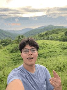a young man wearing glasses takes a selfie in a grassy field with mountains in the background