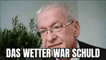 a man wearing glasses and a suit is sitting in front of a plant and a sign that says das wetter war schuld .
