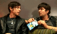 two men are sitting next to each other on a couch and one is holding a bag of chips .
