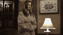 a woman in a trench coat stands in front of a lamp and a framed picture