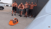 a group of men flexing their muscles in a parking lot with an orange bag in the foreground